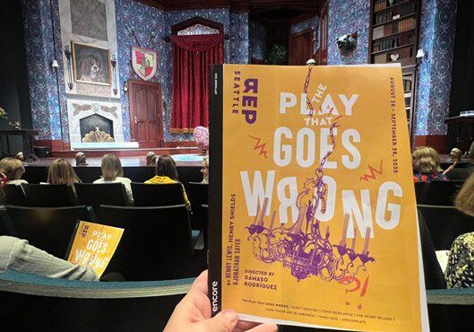 A hand holds a theater program titled 'Play Goes Wrong' inside a theater.