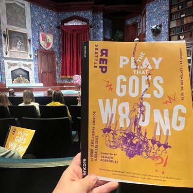 A hand holds a theater program titled 'Play Goes Wrong' inside a theater.